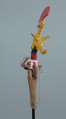Ujęcie z tyłu. Kolorowy ptak zrobiony z drewna z ruchomą żuchwą poruszaną za pomocą sznurka. Długi, pomalowany w kolorowe paski dziób. Na głowie umieszczony drugi, mniejszy ptak w żółtym kolorze z czerwonym czubem.