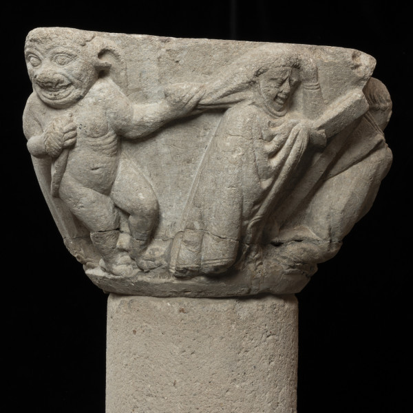 czarci kapitel - Ujęcie zakonnicy ciągnionej za welon przez czarta. Kapitel blokowo-kielichowy, rozszerzający się ku górze, wykuty z jednego kawałka kamienia. Okrągłą podstawę otacza wałek o trójkątnym profilu. Czworoboczne zwieńczenie. Powierzchnia dekorowana płaskorzeźbionymi przedstawieniami pięciu mnichów, stołu eucharystycznego z kielichem i hostią oraz czarta. Postaci o krępej sylwetce i dużych głowach. Brzegi szat dekorowane charakterystycznym podwójnym wałkiem. Czart nagi, o muskularnej budowie, z dużą głową i długimi uszami. Okrągła twarz o migdałowatych oczach, wyszczerzona w szerokim uśmiechu. Relief głęboki, półplastyczny, światłocieniowy. Gładko opracowane tło. Niewielkie obtłuczenia przy podstawie i zwieńczeniu. Brak abakusa. Pęknięcie dookoła kapitela na wysokości powyżej wałka. Pod spodem centralne wgłębienie do umocowania trzpienia łączącego kapitel z trzonem.