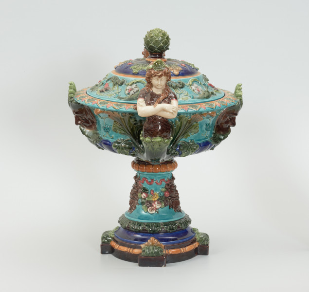 Waza - Ujęcie z lewej strony z pokrywką. Waza w kształcie smukłej amfory. Brzusiec ceramiczny, rozszerzający się ku górze, w kolorze kremowym, malowany podszkliwnie (z uzupełnieniami naszkliwnymi) we wzory kwiatowe - bukiety barwnych kwiatów w typie aster w kolorach fioletowo-czerwonym, liliowym, żółtym i białym, z gałązkami i listkami oraz drobnymi kwiatkami przypominającymi bratki o barwie fioletowo-czerwonej i liliowej. Górna część wazy wraz z uchwytami oraz stopą i część podstawy wykonane jako odlewy z mosiądzu. Mosiężna szyja wąska z otokiem perełkowym w środku ozdobiona jest pionowym, rozszerzającym się ku dołowi i ku górze rytmicznym podziałem z ornamentem geometryczno-roślinnym. Na pogrubionym skraju szyi umieszczona jest płytka miseczka w formie kwiatu o wąskich płatkach, stanowiąca wylewkę wazonu. Poniżej dolnej części szyi oprawa wierzchołka brzuśca składająca się z otoku o sercowym wycięciu zwieńczonym profilowaną palmetką oraz dwóch wydłużonych, sięgających 1/3 wysokości brzuśca, pionowych wsporników uchwytów. Te ostatnie składają się z 2 części: zewnętrzna wychodząca ze wsporników opartych na brzuścu, wyniesiona jest po niewielkim skosie do połowy wysokości szyi i na końcu wywinięta do wnętrza wolutowo, a druga - krótsza, skierowana ku dołowi łączy łagodnym łukiem wolutę z górną częścią oprawy brzuśca, kończąc się wywiniętymi na zewnątrz listkami. Uchwyty żłobkowane, zdobione motywem liści. Wazon wsparty jest na mosiężnej podstawie, składającej się z wieńca oplecionego wstęgą, na którym spoczywa zwężająca się ku górze stopa o brzegach ozdobionych perełkowaniem i pierścieniem, wspierającym koszyczek, ujmujący dolną część ceramicznego brzuśca, w formie kwiatu o ostro zakończonych, wąskich płatkach. Całość spoczywa na podstawie z kwadratowej płytki czerwonego marmuru ze ściętymi narożnikami, ujętymi mosiężnymi wspornikami ramy wspierającej płytę od dołu i opartej w narożach na czterech lwich łapach.