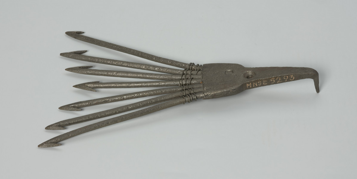 Ość siedmiozębna - Ujęcie przodu z góry. Ość siedmiozębna wykonana z żelaza techniką kowalską. Kształtem przypomina widły z oplecionymi u nasady za pomocą metalowego drutu zębami. Oś narzędzia stanowi najkrótszy ząb, od którego pozostałe żeby rozchodzą się promieniście, przy czym każdy kolejny jest dłuższy od poprzedniego. Zęby zaopatrzone są w ostrza, z których jedynie ostrze środkowego zęba jest obustronne (ma kształt strzałki).  U nasady części pracującej znajduje się szpikulec, który wbijany był trzonek (trzonka brak) oraz okrągły otwór, ułatwiający umocowanie ości do drzewca przy pomocy gwoździa.