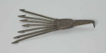 Ość siedmiozębna - Ujęcie przodu z góry. Ość siedmiozębna wykonana z żelaza techniką kowalską. Kształtem przypomina widły z oplecionymi u nasady za pomocą metalowego drutu zębami. Oś narzędzia stanowi najkrótszy ząb, od którego pozostałe żeby rozchodzą się promieniście, przy czym każdy kolejny jest dłuższy od poprzedniego. Zęby zaopatrzone są w ostrza, z których jedynie ostrze środkowego zęba jest obustronne (ma kształt strzałki).  U nasady części pracującej znajduje się szpikulec, który wbijany był trzonek (trzonka brak) oraz okrągły otwór, ułatwiający umocowanie ości do drzewca przy pomocy gwoździa.