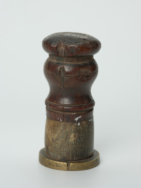 tłok pieczętny - Ujęcie w pionie tyłem pieczęcią w dół. Okragły tłok pieczętny z brązu z drewniano-rogowym uchwytem oraz rysunkiem herbu Białogardu trzymanym przez dzikich ludzi w polu pieczęci.