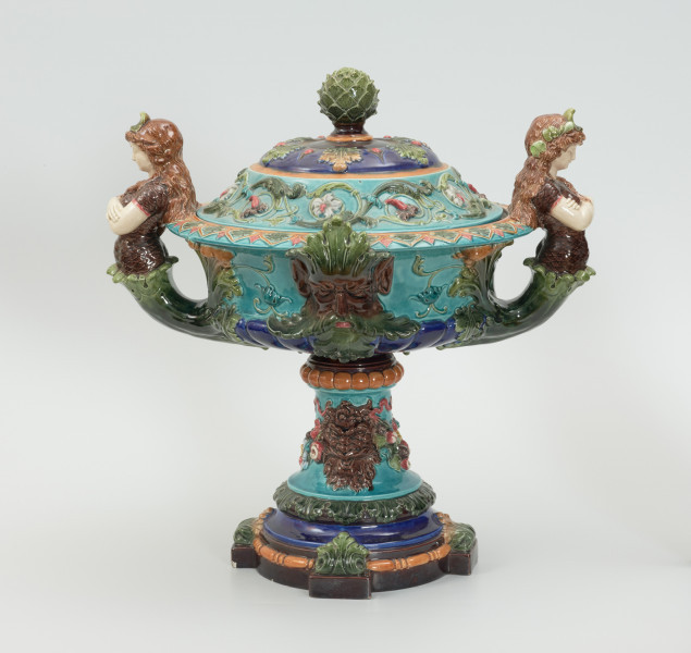 Waza - Ujęcie z przodu z pokrywką. Waza w kształcie smukłej amfory. Brzusiec ceramiczny, rozszerzający się ku górze, w kolorze kremowym, malowany podszkliwnie (z uzupełnieniami naszkliwnymi) we wzory kwiatowe - bukiety barwnych kwiatów w typie aster w kolorach fioletowo-czerwonym, liliowym, żółtym i białym, z gałązkami i listkami oraz drobnymi kwiatkami przypominającymi bratki o barwie fioletowo-czerwonej i liliowej. Górna część wazy wraz z uchwytami oraz stopą i część podstawy wykonane jako odlewy z mosiądzu. Mosiężna szyja wąska z otokiem perełkowym w środku ozdobiona jest pionowym, rozszerzającym się ku dołowi i ku górze rytmicznym podziałem z ornamentem geometryczno-roślinnym. Na pogrubionym skraju szyi umieszczona jest płytka miseczka w formie kwiatu o wąskich płatkach, stanowiąca wylewkę wazonu. Poniżej dolnej części szyi oprawa wierzchołka brzuśca składająca się z otoku o sercowym wycięciu zwieńczonym profilowaną palmetką oraz dwóch wydłużonych, sięgających 1/3 wysokości brzuśca, pionowych wsporników uchwytów. Te ostatnie składają się z 2 części: zewnętrzna wychodząca ze wsporników opartych na brzuścu, wyniesiona jest po niewielkim skosie do połowy wysokości szyi i na końcu wywinięta do wnętrza wolutowo, a druga - krótsza, skierowana ku dołowi łączy łagodnym łukiem wolutę z górną częścią oprawy brzuśca, kończąc się wywiniętymi na zewnątrz listkami. Uchwyty żłobkowane, zdobione motywem liści. Wazon wsparty jest na mosiężnej podstawie, składającej się z wieńca oplecionego wstęgą, na którym spoczywa zwężająca się ku górze stopa o brzegach ozdobionych perełkowaniem i pierścieniem, wspierającym koszyczek, ujmujący dolną część ceramicznego brzuśca, w formie kwiatu o ostro zakończonych, wąskich płatkach. Całość spoczywa na podstawie z kwadratowej płytki czerwonego marmuru ze ściętymi narożnikami, ujętymi mosiężnymi wspornikami ramy wspierającej płytę od dołu i opartej w narożach na czterech lwich łapach.