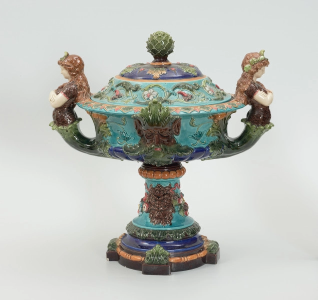 Waza - Ujęcie z przodu z pokrywką. Waza w kształcie smukłej amfory. Brzusiec ceramiczny, rozszerzający się ku górze, w kolorze kremowym, malowany podszkliwnie (z uzupełnieniami naszkliwnymi) we wzory kwiatowe - bukiety barwnych kwiatów w typie aster w kolorach fioletowo-czerwonym, liliowym, żółtym i białym, z gałązkami i listkami oraz drobnymi kwiatkami przypominającymi bratki o barwie fioletowo-czerwonej i liliowej. Górna część wazy wraz z uchwytami oraz stopą i część podstawy wykonane jako odlewy z mosiądzu. Mosiężna szyja wąska z otokiem perełkowym w środku ozdobiona jest pionowym, rozszerzającym się ku dołowi i ku górze rytmicznym podziałem z ornamentem geometryczno-roślinnym. Na pogrubionym skraju szyi umieszczona jest płytka miseczka w formie kwiatu o wąskich płatkach, stanowiąca wylewkę wazonu. Poniżej dolnej części szyi oprawa wierzchołka brzuśca składająca się z otoku o sercowym wycięciu zwieńczonym profilowaną palmetką oraz dwóch wydłużonych, sięgających 1/3 wysokości brzuśca, pionowych wsporników uchwytów. Te ostatnie składają się z 2 części: zewnętrzna wychodząca ze wsporników opartych na brzuścu, wyniesiona jest po niewielkim skosie do połowy wysokości szyi i na końcu wywinięta do wnętrza wolutowo, a druga - krótsza, skierowana ku dołowi łączy łagodnym łukiem wolutę z górną częścią oprawy brzuśca, kończąc się wywiniętymi na zewnątrz listkami. Uchwyty żłobkowane, zdobione motywem liści. Wazon wsparty jest na mosiężnej podstawie, składającej się z wieńca oplecionego wstęgą, na którym spoczywa zwężająca się ku górze stopa o brzegach ozdobionych perełkowaniem i pierścieniem, wspierającym koszyczek, ujmujący dolną część ceramicznego brzuśca, w formie kwiatu o ostro zakończonych, wąskich płatkach. Całość spoczywa na podstawie z kwadratowej płytki czerwonego marmuru ze ściętymi narożnikami, ujętymi mosiężnymi wspornikami ramy wspierającej płytę od dołu i opartej w narożach na czterech lwich łapach.