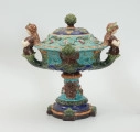 Waza - Ujęcie z przodu z pokrywką. Waza w kształcie smukłej amfory. Brzusiec ceramiczny, rozszerzający się ku górze, w kolorze kremowym, malowany podszkliwnie (z uzupełnieniami naszkliwnymi) we wzory kwiatowe - bukiety barwnych kwiatów w typie aster w kolorach fioletowo-czerwonym, liliowym, żółtym i białym, z gałązkami i listkami oraz drobnymi kwiatkami przypominającymi bratki o barwie fioletowo-czerwonej i liliowej. Górna część wazy wraz z uchwytami oraz stopą i część podstawy wykonane jako odlewy z mosiądzu. Mosiężna szyja wąska z otokiem perełkowym w środku ozdobiona jest pionowym, rozszerzającym się ku dołowi i ku górze rytmicznym podziałem z ornamentem geometryczno-roślinnym. Na pogrubionym skraju szyi umieszczona jest płytka miseczka w formie kwiatu o wąskich płatkach, stanowiąca wylewkę wazonu. Poniżej dolnej części szyi oprawa wierzchołka brzuśca składająca się z otoku o sercowym wycięciu zwieńczonym profilowaną palmetką oraz dwóch wydłużonych, sięgających 1/3 wysokości brzuśca, pionowych wsporników uchwytów. Te ostatnie składają się z 2 części: zewnętrzna wychodząca ze wsporników opartych na brzuścu, wyniesiona jest po niewielkim skosie do połowy wysokości szyi i na końcu wywinięta do wnętrza wolutowo, a druga - krótsza, skierowana ku dołowi łączy łagodnym łukiem wolutę z górną częścią oprawy brzuśca, kończąc się wywiniętymi na zewnątrz listkami. Uchwyty żłobkowane, zdobione motywem liści. Wazon wsparty jest na mosiężnej podstawie, składającej się z wieńca oplecionego wstęgą, na którym spoczywa zwężająca się ku górze stopa o brzegach ozdobionych perełkowaniem i pierścieniem, wspierającym koszyczek, ujmujący dolną część ceramicznego brzuśca, w formie kwiatu o ostro zakończonych, wąskich płatkach. Całość spoczywa na podstawie z kwadratowej płytki czerwonego marmuru ze ściętymi narożnikami, ujętymi mosiężnymi wspornikami ramy wspierającej płytę od dołu i opartej w narożach na czterech lwich łapach.