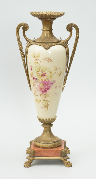 Waza - Ujęcie z tyłu.  Waza dekoracyjna o bogatej, barwnej dekoracji reliefowej, w kształcie kielicha ze spłaszczoną czarą z dwoma uchwytami i przykrywą. Podstawa okrągła, profilowana z czterema prostokątnymi poszerzeniami z listkiem akantu. Trzon na styku z podstawą ozdobiony wałkiem z liści akantu, w części środkowej wypełniony reliefem z dwiema brązowymi maskami i girlandami drobnych, barwnych kwiatów. W górnej części trzonu perełkowy otok z przewężeniem na styku z czarą. Czara z szerokim brzegiem ozdobionym dekoracją geometryczną i zwężającym się do dołu brzuścem dekorowanym dwiema wypełniającymi całą wysokość czary brązowymi maskami, których zielone grzywy w formie palmety opierają się na górnej krawędzi brzegu, a brody na spodzie czary ozdobionym granatowymi 