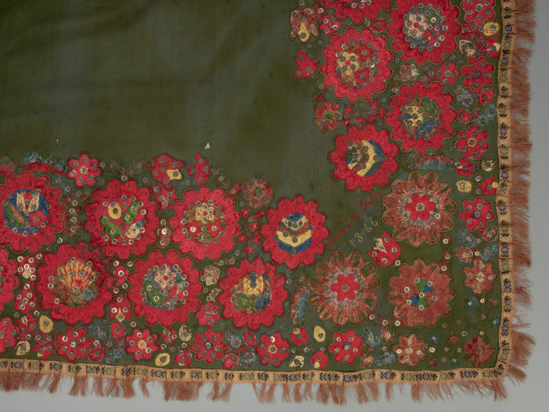 zielona chusta z, haftem z motywami kwiatowymi, cekinami i frędzlami - detal; Fragment prawego górnego rogu chusty. Haft płaski z motywem wielopłatkowych kwiatków w kolorze czerwonym na zielonym materiale. Środki kwiatów o kolorach niebieskim, żółtym i zielonym układają się w różne wzory florystyczne. Każdy z kwiatów obwiedziony jest złotym sznureczkiem. Pomiędzy większymi dodatkowo wyhaftowane są drobniejsze kwiatki. całość zdobiona także cekinami. Pomiędzy kwiatami w rogu napis: 