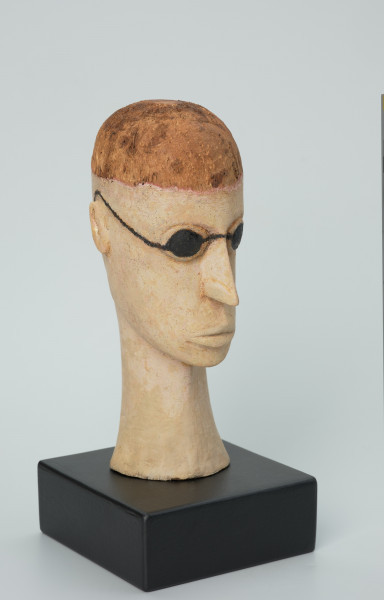 lalka teatralna: Tubabu Cè Kun - Ujęcie z przodu z prawego boku. Drewniane popiersie mężczyzny.  Głowa owalna, wydłużona, zaznaczona linia włosów nad czołem. Włosy pomalowane na kolor jasno-brązowy/blond. Nos długi, wąski, usta wypukle, pomalowane na kolor jasno-różowy. Zamiast oczu czarne okulary, okrągłe, z cienkimi nausznicami. Spod okularów wystające powieki dolne i górne. Szyja długa, wąska, rozszerzona u dołu, pomalowana na kolor jasno-różowy.