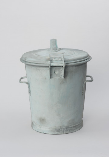 metalowy pojemnik z pokrywą - Ujęcie z tyłu; Metalowy pojemnik z pokrywą służący do przechowywania odpadków. Pojemnik zaopatrzony w dwa boczne uchwyty do transportu ręcznego oraz zaczep służący prawdopodobnie do przewozu pojemnika na specjalnym wózku transportowym. Klapa uchylna połączona zawiasem z korpusem pojemnika.