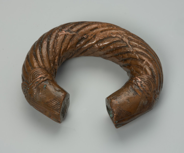 biżuteria, bransoleta - Ujęcie z przodu z dołu. Otwarta, miedziana, owalna bransoleta ozdobiona grubymi, zaokrąglonymi, biegnącymi dookolnie prążkami; cienkimi, ukośnymi nacięciami oraz półkolami przypominającymi wachlarze wyłaniające się z motywu plecionki.