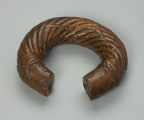 biżuteria, bransoleta - Ujęcie z przodu z dołu. Otwarta, miedziana, owalna bransoleta ozdobiona grubymi, zaokrąglonymi, biegnącymi dookolnie prążkami; cienkimi, ukośnymi nacięciami oraz półkolami przypominającymi wachlarze wyłaniające się z motywu plecionki.