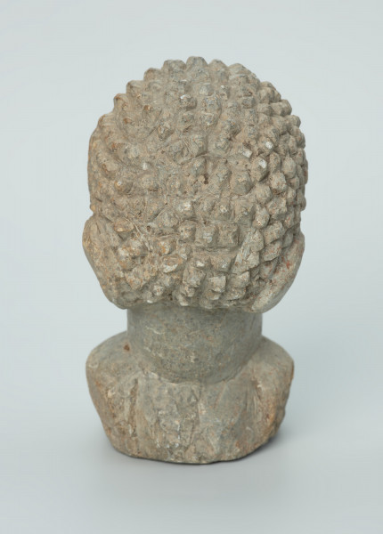 rzeźba; głowa; figura kultu zmarłych - Ujęcie z tyłu. Rzeźbiona w szarobeżowym steatycie, usadowiona na okrągłej podstawce, głowa postaci ludzkiej z dużymi uszami z tyłu i w charakterystycznym uczesani w formie małych, lekko zaokrąglonych stożków, być może loczków.
