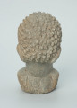 rzeźba; głowa; figura kultu zmarłych - Ujęcie z tyłu. Rzeźbiona w szarobeżowym steatycie, usadowiona na okrągłej podstawce, głowa postaci ludzkiej z dużymi uszami z tyłu i w charakterystycznym uczesani w formie małych, lekko zaokrąglonych stożków, być może loczków.