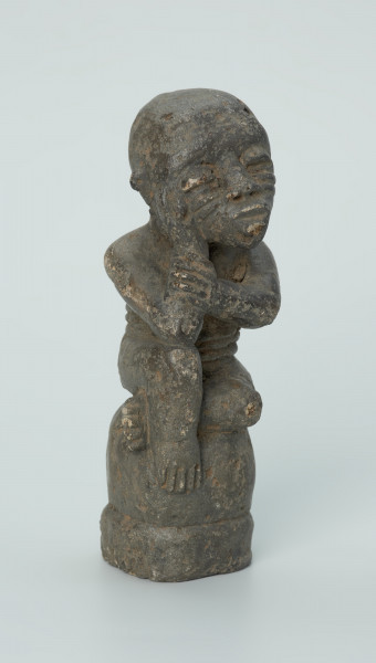 figura kultu przodków - Ujęcie z przodu z prawej. Rzeźbiona w grafitowym kamieniu postać ludzka w pozycji siedzącej - najprawdopodobniej mężczyzny. Postać siedzi na wysokiej podstawie, która swym kształtem przypomina kamień. Rzeźba ma charakterystyczne poziome nacięcia widoczne na obu policzkach. Widoczne rysy, mikropęknięcia i liczne malutkie otworki.