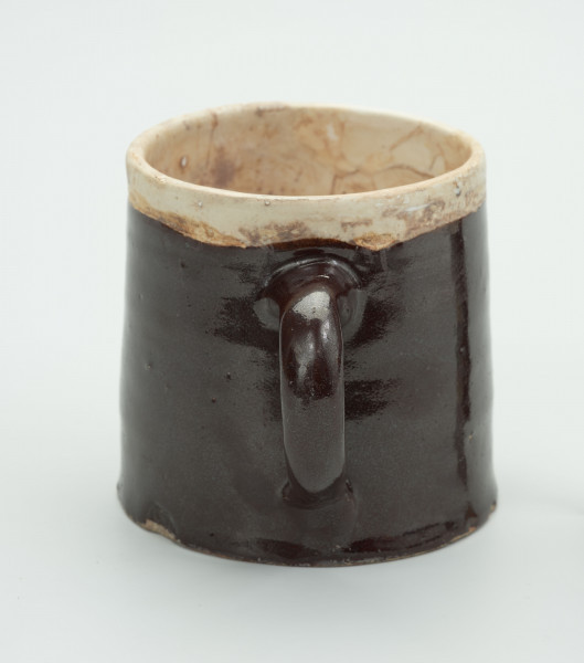 Kamionkowy kubek w kształcie kufla - Ujęcie z tyłu; Kubek w kształcie walca zwężającego się ku górze. Wlew węższy od dna. Szkliwo dwukolorowe; wokół wylewu i w środku jasnobeżowe/kremowe, pozostała część i ucho w kolorze ciemnego brązu. Ucho wzdłużne, taśmowe umieszczone od linii jasnego szkliwa i zakończone na 4/5 wysokości naczynia.