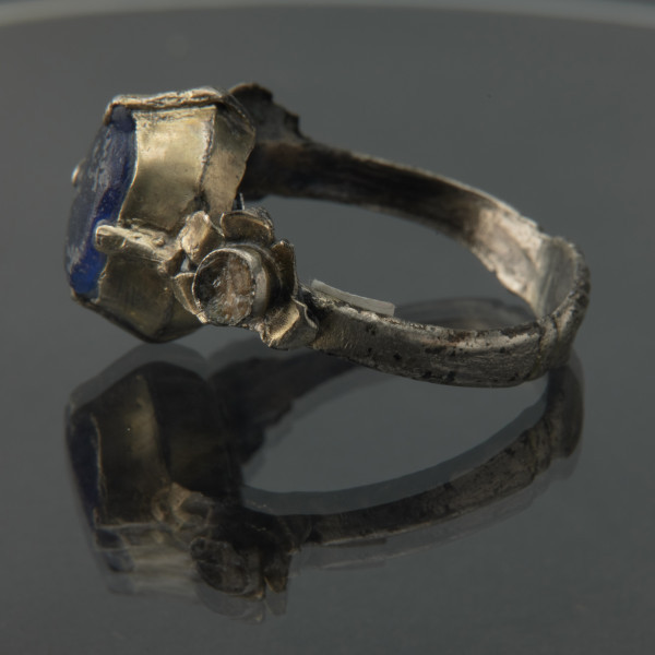 Pozłacany pierścień z niebieskim oczkiem - Ujęcie prawej strony. Pierścień z niebieskim oczkiem.  Oczko szklane, niebieskie, płaskie i owalne.. Z boku dwie mniejsza owalna oprawa, osadzona w pięciopłatkowej rozecie. W oprawce oczko.