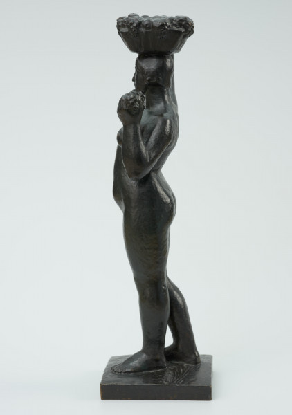 akt kobiecy - Ujęcie z boku z lewej strony; Posążek przedstawia postać stojącej kobiety o pełnych kształtach dźwigającą kosz na głowie. Sylwetka wyprostowana o masywnych nogach, biodrach i niewielkich piersiach, zupełnie naga, z okrągłą twarzą i krótkich włosach- prawa noga lekko cofnięta, lewa ręka zgięta w łokciu dzierży na wysokości barku kiść owoców, prawa uniesiona ręka przytrzymuje na głowie płaski okrągły niski kosz wypełniony owocami. Figurka stoi na kwadratowej niskiej podstawce.