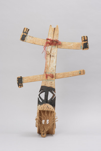 maska kanaga - kompletna - Ujęcie z tyłu. Maska wykonana z materiałów organicznych: skóry, włókna roślinnego i drewna. Pionowa część w górnej połowie wypełniona białymi znakami, dolna część pomalowana na czarno. Do części pionowej przymocowane dwie listwy poziome również pomalowane w białe znaki.