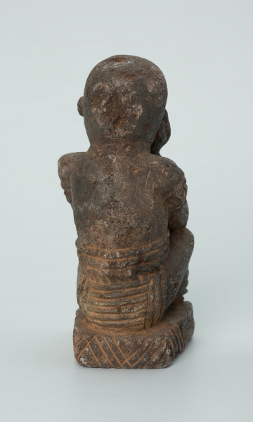 Ujęcie z tyłu. Rzeźbiona w grafitowym kamieniu postać ludzka - najprawdopodobniej mężczyzna - w pozycji siedzącej z nogami skrzyżowanymi. Rzeźba ma charakterystyczne bransolety – dwie na ramionach oraz jedną na nadgarstku - które ozdobiono wzorem w postaci ukośnych linii. Widoczne ślady czerwonego piasku, rysy, mikropęknięcia oraz ubytek z tyłu głowy.