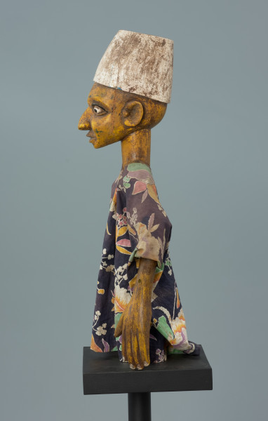 lalka teatralna: Kalhaldi (muzułmański uczony) - Ujęcie z lewego boku. Lalka teatralna przedstawiająca postać męską w nakryciu głowy w formie walca rozszerzonego u dołu, pomalowanego na biało. Szata z kwiecistego materiału, szeroka, z szerokimi rękawami. Oczy białe, z czarnymi źrenicami, drobne i wypukłe, z zaznaczonymi powiekami dolnymi oraz łukowatymi czarnymi brwiami.  Nos rozszerzony u dołu, usta zamknięte, z dolną wargą pełniejszą. Ręce przy tułowiu. Dłonie z zaznaczonymi palcami.