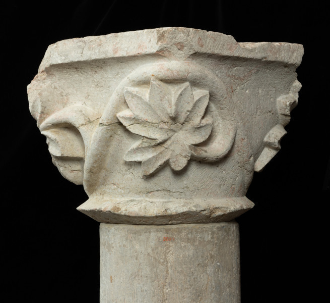 Kapitel z przedstawieniem łodygi passiflory - Ujęcie drugiego boku. Kapitel blokowo-kielichowy, rozszerzający się ku górze, wykuty z jednego kawałka kamienia. Okrągłą podstawę otacza wałek o trójkątnym profilu. Zwieńczenie w formie szerokiej czworobocznej płyty abakusa. Pomiędzy wałkiem i abakusem płaskorzeźbiona, wijąca się dookoła łodyga passiflory. Z niej wyrastają rozłożyste liście o palmetowym kształcie, akcentujące narożniki kapitela. Forma rośliny pełna, mięsista. Tło opracowane gładko. Drobne pęknięcia na powierzchni kamienia. Narożnik abakusa ukruszony. Pod spodem centralne wgłębienie do umocowania trzpienia łączącego kapitel z trzonem.