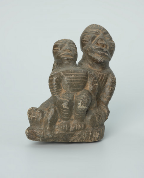 figura kultu przodków - Ujęcie z przodu. Rzeźbione w grafitowym kamieniu dwie postaci ludzkie w pozycji siedzącej. Mniejsza postać – najprawdopodobniej dziecko - siedzi na kolanach większej - najprawdopodobniej kobiety. Obie postacie ozdobiono licznymi nacięciami liniowymi. Widoczne rysy i mikropęknięcia.