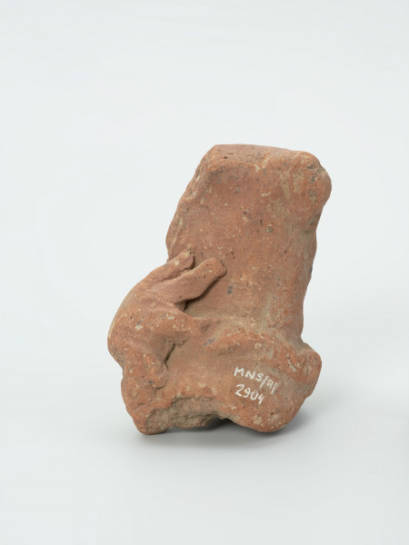 głowa - Ujęcie z tyłu; Gliniana głowa postaci ludzkiej o ceglastym kolorze i prostokątnej twarzy, która opiera się na masywnej szyi. Zachowało się lewe ramię, na którym wymodelowano charakterystyczną ozdobę w kształcie koła, od którego odchodzą promienie. Figurce zaznaczono rysy twarzy: wydatne, rozchylone usta, szeroki nos o okrągłych nozdrzach oraz wypukłe, okrągłe oczy, które przypominają dwie kulki gliny. Na głowie postaci zaznaczono dwa okrągłe zgrubienia (guzki), które prawdopodobnie przedstawiają fryzurę lub nakrycie głowy. Na powierzchni widoczne są rysy, mikropęknięcia oraz przetarcia.