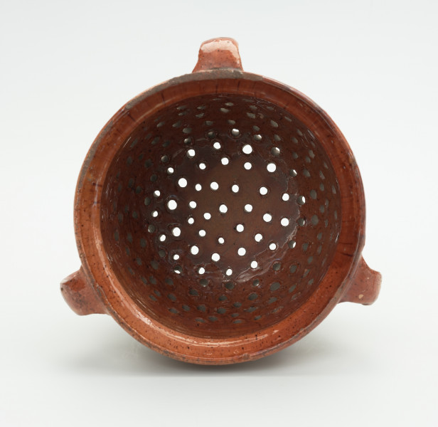 Gliniany durszlak z trzema uchami - Ujęcie wnętrza. Gliniane, okrągłe naczynie z widocznymi trzema uszkami. Wewnatrz z dzirkami rozmieszczonymi nieregularnie.