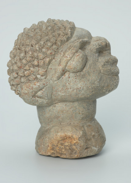 rzeźba; głowa; figura kultu zmarłych - Ujęcie prawy bok. Rzeźbiona w szarobeżowym steatycie, usadowiona na okrągłej podstawce, głowa postaci ludzkiej z dużymi uszami z tyłu i w charakterystycznym uczesani w formie małych, lekko zaokrąglonych stożków, być może loczków.