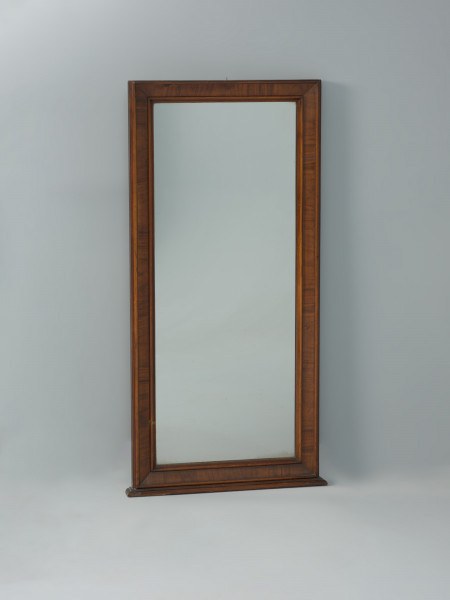 Szklane lustro z fornirowaną ramą - Ujęcie z przodu. Lustro z jednej tafli szkła, oprawione w drewnianą, fornirowaną ramę, w której listwowy profil zewnętrzny i wewnętrzny ujmuje szerszą część środkową ustawioną po skosie, tworzącym zagłębienie ramy. Wąska profilowana podstawa z kołkami do osadzenia na szafce.