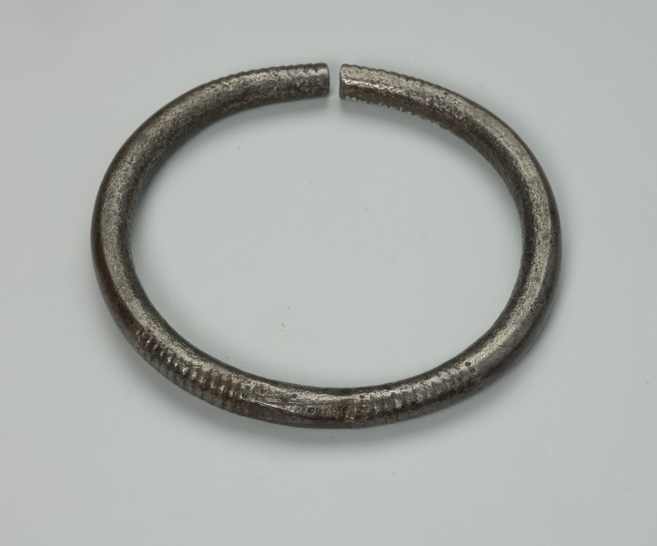 biżuteria, bransoleta - Ujęcie z tyłu z dołu. Otwarta, żelazna, okrągła bransoleta ozdobiona nacięciami w postaci pionowych linii oraz wgłębieniami w kształcie maleńkich kropek. Zamknięcie bransolety w formie szpary.