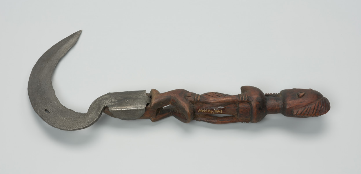 sierp - Ujęcie z przodu w poziomie. Sierp o kutym z żelaza, łukowatym ostrzu z drewnianą rączką w formie rzeźbionej stojącej postaci z biustem i brodą z rękoma na udach.