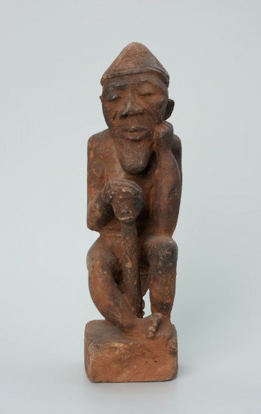 figura kultu przodków - Ujęcie z przodu. Rzeźbiona w brunatno-czerwonym kamieniu dość duża postać mężczyzny (starca?) w pozycji siedzącej. Mężczyzna siedzi na okrągłym, malutkim stołku, ma charakterystyczne nakrycie głowy - spiczastą czapkę o zaokrąglonych rogach. Jego prawa ręka opiera się o berło (laskę) zakończone rzeźbą głowy. Widoczne rysy, mikropęknięcia i liczne malutkie otworki.