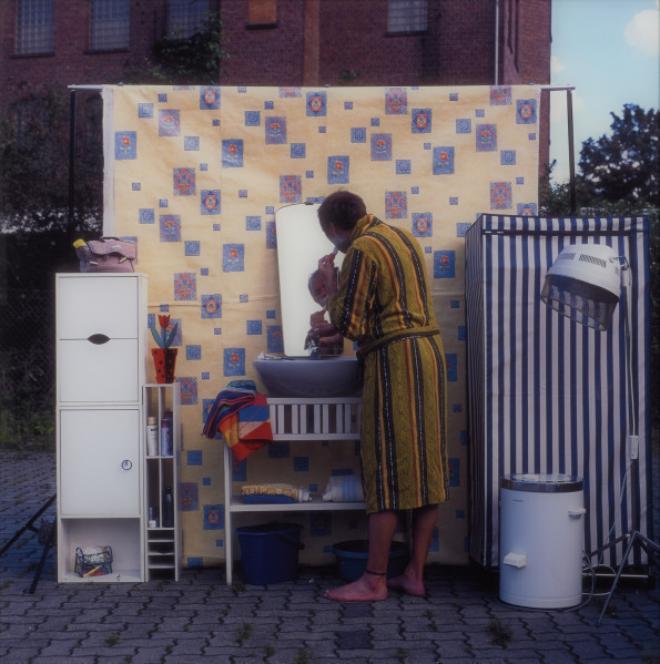 fotografia - Ujęcie z przodu. Fotografia barwna przedstawiająca mężczyznę golącego się w zaaranżowanej w przestrzeni miejskiej łazience. W tle elewacja ceglanego budynku.