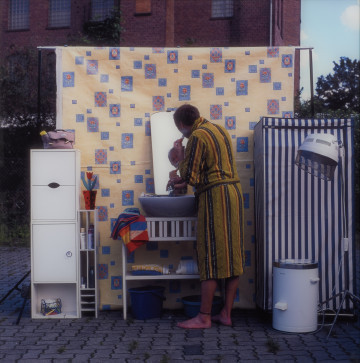 fotografia - Ujęcie z przodu. Fotografia barwna przedstawiająca mężczyznę golącego się w zaaranżowanej w przestrzeni miejskiej łazience. W tle elewacja ceglanego budynku.