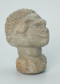 rzeźba; głowa; figura kultu zmarłych - Ujęcie z przodu z prawej strony. Rzeźbiona w szarobeżowym steatycie, usadowiona na okrągłej podstawce, głowa postaci ludzkiej z dużymi uszami z tyłu i w charakterystycznym uczesani w formie małych, lekko zaokrąglonych stożków, być może loczków.