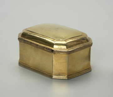 pozłacane puzderko na niskim, profilowanym cokoliku - Ujęcie z tyłu, z ukosa; Pozłacane puzderko na niskim, profilowanym cokoliku. Korpus ośmioboczny, na rzucie prostokąta ze ściętymi narożami. Pokrywa na zawiasie, z dwoma uskokami, lekko wybrzuszona z wielokrotnie profilowanym brzegiem, z przodu w centralnej części wysunięty wydłużony płaski uchwyt w formie listwy o zaoblonych narożach, trójkątnym wykroju i z delikatnym profilowaniem. Powierzchnia puzderka na zewnątrz i we wnętrzu złocona, poza częścią spodnią.