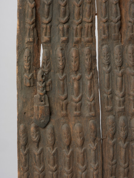 drzwi - Ujęcie z przodu z prawej strony w przybliżeniu. Drzwi drewniane do spichlerza, składające się z dwóch części połączonych klamerkami z żelaznej blachy. Zdobione płaskorzeźbionymi postaciami o schematycznych rysach z dłońmi na kolanach. Postaci w pięciu poziomych rzędach po 12 i 13 w rzędzie. Na wysokości czwartego rzędu przymocowana z lewej strony nieruchoma część zasuwy, wykonana z drewna i zdobiona rzeźbioną siedzącą postacią. Ruchomej części zasuwy brak.