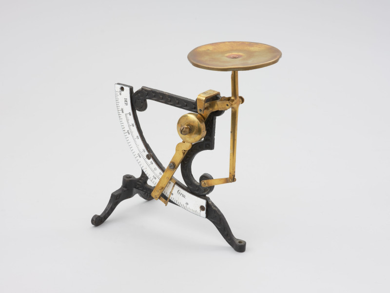 jednowahadłowa uchylna waga pocztowa - Ujęcie z przodu lewoboczne; Jednowahadłowa uchylna waga pocztowa. Waga składa się ze stalowej skali o wartości od 0 do 350 gram, z podziałką co 5 gram, która połączona jest z trzonem wagi za pomocą stalowych blaszek znitowanych na ruchomo. Podstawa wagi składa się z dwóch elementów, z których element poprzeczny przymocowany jest do głównego elementu na ruchomo, co pozwala składać nóżkę poprzez jej przekręcenie. Dłuższe ramię podstawy zakończone jest śrubką pozwalającą na regulację wagi w opozycji pionowej. Stalowa konstrukcja łącząca główny trzon wagi z dźwignią nośną nasadzona jest na szalkę.