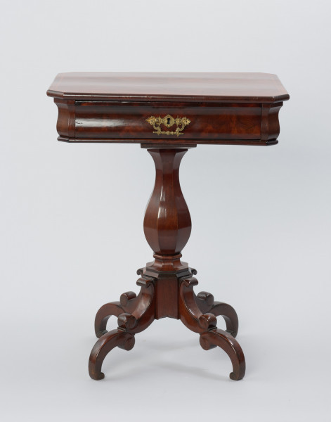 Stolik do robótek - Ujęcie z przodu. Stolik z prostokątnym blatem ze ściętymi narożami i sfazowaną krawędzią, ozdobiony intarsją w formie wąskiego otoku z geometrycznym wzorem. Ścianka pod blatem o wybrzuszonej linii przekroju kryje szufladę z mosiężnym okuciem z dziurką do klucza i ruchomym uchwytem w centrum ścianki czołowej. Stolik wsparty na nodze w formie wydłużonej ośmiobocznej tralki gruszkowej, opartej przewężonym profilem na ośmiobocznym trzonie, do którego przymocowane są po przekątnej cztery nóżki składające się z dwóch odwróconych i połączonych esownic. Wewnątrz stolika znajduje się szuflada, która podzielona jest na jedenaście symetrycznie rozmieszczonych przegródek różnej wielkości, z krawędziami podmalowanymi na czarno.