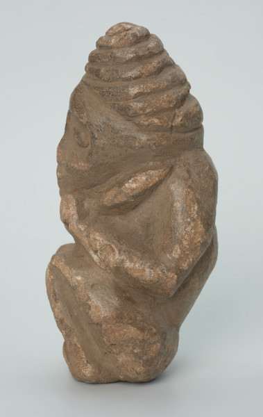 rzeźba; przedmiot obrzędowy; Figura kultu zmarłych - Ujęcie lewy bok. Rzeźbiona w szarobeżowym steatycie postać ludzka ze spiralną linią na czubku dużej głowy, siedząca z przykurczonymi nogami i trzymająca w dłoniach bliżej nieokreślone przedmioty, kształtem przypominające motyki.