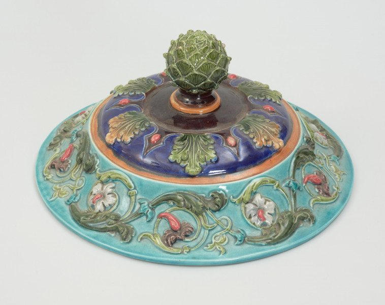 Waza - Ujęcie pokrywki z góry. Waza w kształcie smukłej amfory. Brzusiec ceramiczny, rozszerzający się ku górze, w kolorze kremowym, malowany podszkliwnie (z uzupełnieniami naszkliwnymi) we wzory kwiatowe - bukiety barwnych kwiatów w typie aster w kolorach fioletowo-czerwonym, liliowym, żółtym i białym, z gałązkami i listkami oraz drobnymi kwiatkami przypominającymi bratki o barwie fioletowo-czerwonej i liliowej. Górna część wazy wraz z uchwytami oraz stopą i część podstawy wykonane jako odlewy z mosiądzu. Mosiężna szyja wąska z otokiem perełkowym w środku ozdobiona jest pionowym, rozszerzającym się ku dołowi i ku górze rytmicznym podziałem z ornamentem geometryczno-roślinnym. Na pogrubionym skraju szyi umieszczona jest płytka miseczka w formie kwiatu o wąskich płatkach, stanowiąca wylewkę wazonu. Poniżej dolnej części szyi oprawa wierzchołka brzuśca składająca się z otoku o sercowym wycięciu zwieńczonym profilowaną palmetką oraz dwóch wydłużonych, sięgających 1/3 wysokości brzuśca, pionowych wsporników uchwytów. Te ostatnie składają się z 2 części: zewnętrzna wychodząca ze wsporników opartych na brzuścu, wyniesiona jest po niewielkim skosie do połowy wysokości szyi i na końcu wywinięta do wnętrza wolutowo, a druga - krótsza, skierowana ku dołowi łączy łagodnym łukiem wolutę z górną częścią oprawy brzuśca, kończąc się wywiniętymi na zewnątrz listkami. Uchwyty żłobkowane, zdobione motywem liści. Wazon wsparty jest na mosiężnej podstawie, składającej się z wieńca oplecionego wstęgą, na którym spoczywa zwężająca się ku górze stopa o brzegach ozdobionych perełkowaniem i pierścieniem, wspierającym koszyczek, ujmujący dolną część ceramicznego brzuśca, w formie kwiatu o ostro zakończonych, wąskich płatkach. Całość spoczywa na podstawie z kwadratowej płytki czerwonego marmuru ze ściętymi narożnikami, ujętymi mosiężnymi wspornikami ramy wspierającej płytę od dołu i opartej w narożach na czterech lwich łapach.