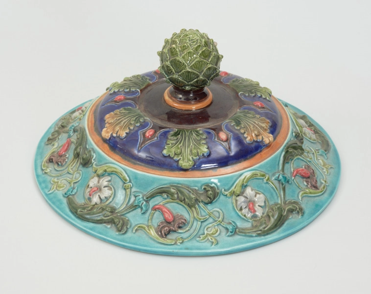 Waza - Ujęcie pokrywki z góry. Waza w kształcie smukłej amfory. Brzusiec ceramiczny, rozszerzający się ku górze, w kolorze kremowym, malowany podszkliwnie (z uzupełnieniami naszkliwnymi) we wzory kwiatowe - bukiety barwnych kwiatów w typie aster w kolorach fioletowo-czerwonym, liliowym, żółtym i białym, z gałązkami i listkami oraz drobnymi kwiatkami przypominającymi bratki o barwie fioletowo-czerwonej i liliowej. Górna część wazy wraz z uchwytami oraz stopą i część podstawy wykonane jako odlewy z mosiądzu. Mosiężna szyja wąska z otokiem perełkowym w środku ozdobiona jest pionowym, rozszerzającym się ku dołowi i ku górze rytmicznym podziałem z ornamentem geometryczno-roślinnym. Na pogrubionym skraju szyi umieszczona jest płytka miseczka w formie kwiatu o wąskich płatkach, stanowiąca wylewkę wazonu. Poniżej dolnej części szyi oprawa wierzchołka brzuśca składająca się z otoku o sercowym wycięciu zwieńczonym profilowaną palmetką oraz dwóch wydłużonych, sięgających 1/3 wysokości brzuśca, pionowych wsporników uchwytów. Te ostatnie składają się z 2 części: zewnętrzna wychodząca ze wsporników opartych na brzuścu, wyniesiona jest po niewielkim skosie do połowy wysokości szyi i na końcu wywinięta do wnętrza wolutowo, a druga - krótsza, skierowana ku dołowi łączy łagodnym łukiem wolutę z górną częścią oprawy brzuśca, kończąc się wywiniętymi na zewnątrz listkami. Uchwyty żłobkowane, zdobione motywem liści. Wazon wsparty jest na mosiężnej podstawie, składającej się z wieńca oplecionego wstęgą, na którym spoczywa zwężająca się ku górze stopa o brzegach ozdobionych perełkowaniem i pierścieniem, wspierającym koszyczek, ujmujący dolną część ceramicznego brzuśca, w formie kwiatu o ostro zakończonych, wąskich płatkach. Całość spoczywa na podstawie z kwadratowej płytki czerwonego marmuru ze ściętymi narożnikami, ujętymi mosiężnymi wspornikami ramy wspierającej płytę od dołu i opartej w narożach na czterech lwich łapach.