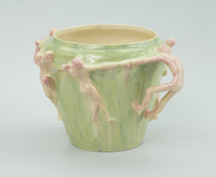 ceramika - Ujęcie z boku; Ceramiczne postacie przyczepione do malowanego wazonu. Postacie mężczyzn i kobiet ciągnące się za ręce. Na przodzie postać prawdopodobnie żeńska zwrócona tyłem do widza. Postać z prawej ciągnie kobietę za rękę zapierając się na stopach i głowie z wygiętym do tyłu tułowiem. Wazon malowany w zielonych barwach. Postacie kremowe z domieszką czerwonej barwy.