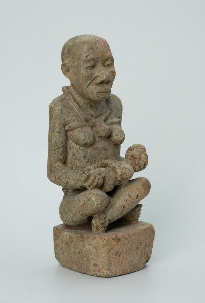 figura kultu przodków - Ujęcie skosem z prawej.  Rzeźbiona w biało-szarym kamieniu dość duża postać kobiety w pozycji siedzącej z nogami skrzyżowanymi. Kobieta siedzi na małej, kwadratowej, lekko zaokrąglonej na rogach podstawie, w dłoniach trzyma dziecko. Widoczne rysy, mikropęknięcia i liczne malutkie otworki.