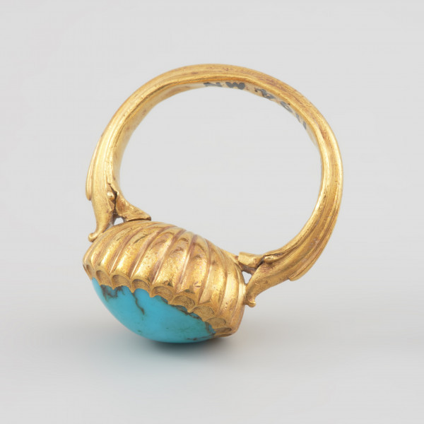 Pierścień z turkusem księcia Franciszka I (1577 - 1620) - Ujęcie z góry. Pierścień z dużym owalnym turkusem w promieniście puklowanej pełnej oprawie. Szyna o profilowanych brzegach, rozszerzająca się w stronę oprawy. Końce szyny ujmujące oprawę kamienia zakomponowane zostały z motywów roślinnych, zbliżonych kształtem do paszczy fantastycznych zwierząt.