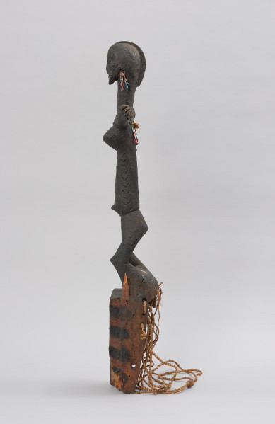 rzeźbiona maska - Ujęcie lewego boku. Drewniana maska z dużymi otworami na oczy, do której przymocowany jest bawełniany sznurek. Maska przyozdobiona jest wyrzeźbioną postacią kobiety z podniesionymi rękami.