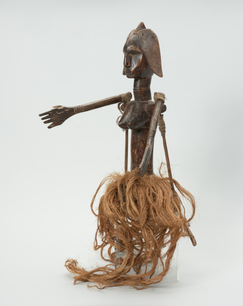 Ujęcie z przodu z lewej strony. Lalka umieszczona na koszyku-czapeczce pokrytej włóknami roślinnymi, noszona na głowie, przedstawiająca kobietę od pasa w górę. Linia brwi i nos wyodrębnione z twarzy. Usta lekko zaznaczone. Twarz ozdobiona skaryfikacją: po dwie linie pionowe na czole i policzkach. Ręce ruchome, poruszają się w linii poziomej za pomocą patyków umocowanych w 