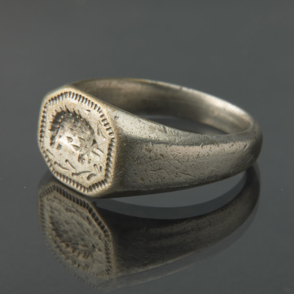 Pozłacany sygnet z wizerunkiem jeża - Ujęcie z przodu skosem w lewą stronę. Srebrny szeroki pierścień z tarczą. Szyna w przekroju półkolista, przy oczku szeroka, profilowana wielobocznie. Oczko w formie sześciokąta, W centrum przedstawienie jeża, wykonane wklęsłym rytem.