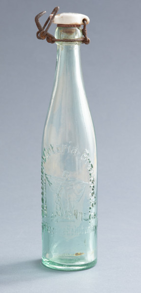 szklana butelka z kapslem - Ujęcie z przodu; Butelka piwówka z grubego szkła przeźroczystego półbiałego z odcieniem zielonym. Szyjka przechodząca łagodnie z korpusu zakończona przegrubieniem z wklęską na drut zamknięcia bocznego - dźwigniowego. Dno wklęsłe. Kapsel z białego porcelitu owalny z wypustami na dwóch bokach.